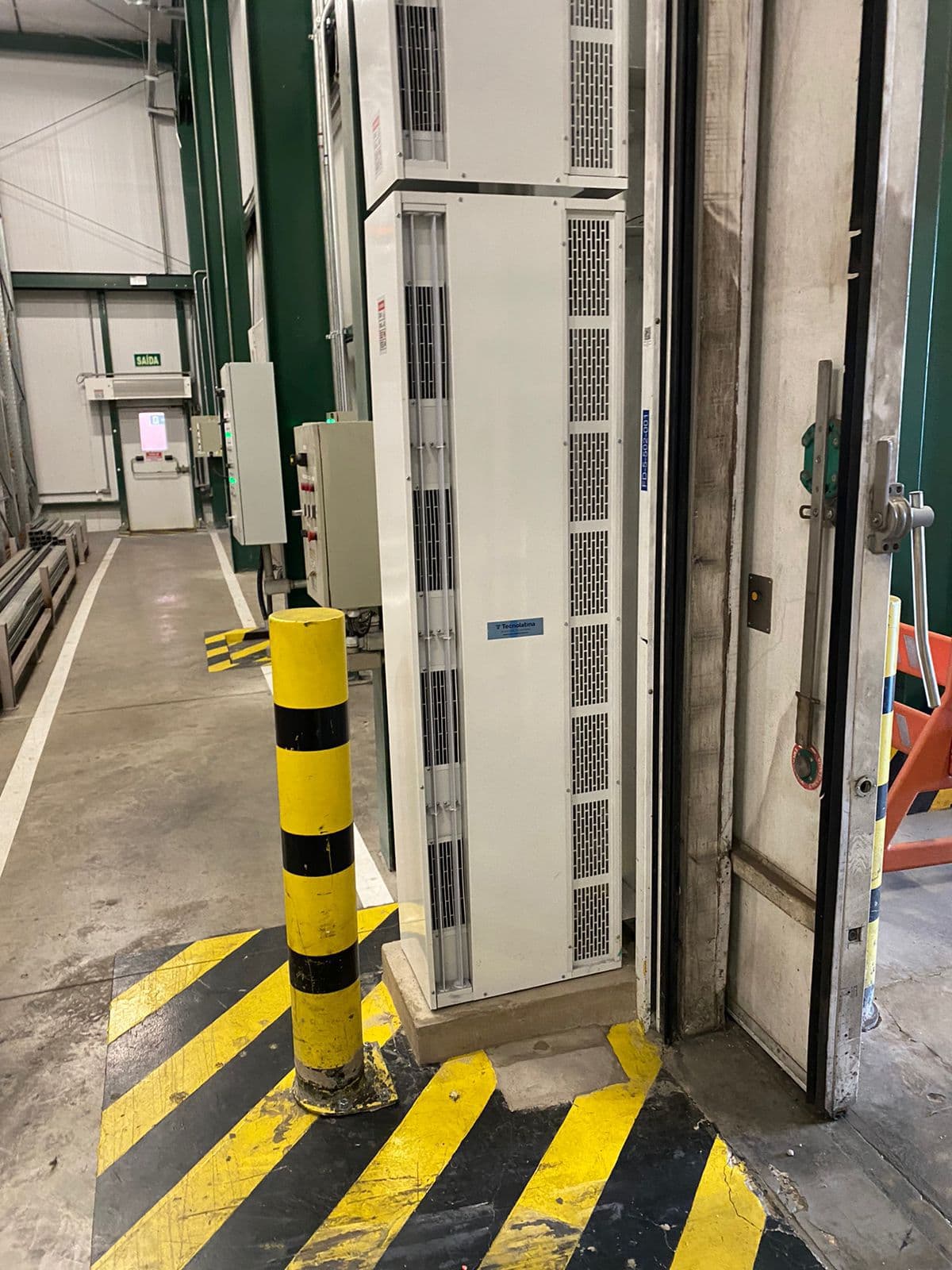 Cortina de ar vertical instalada ao lado de porta industrial, junto a proteção de piso amarela e preta